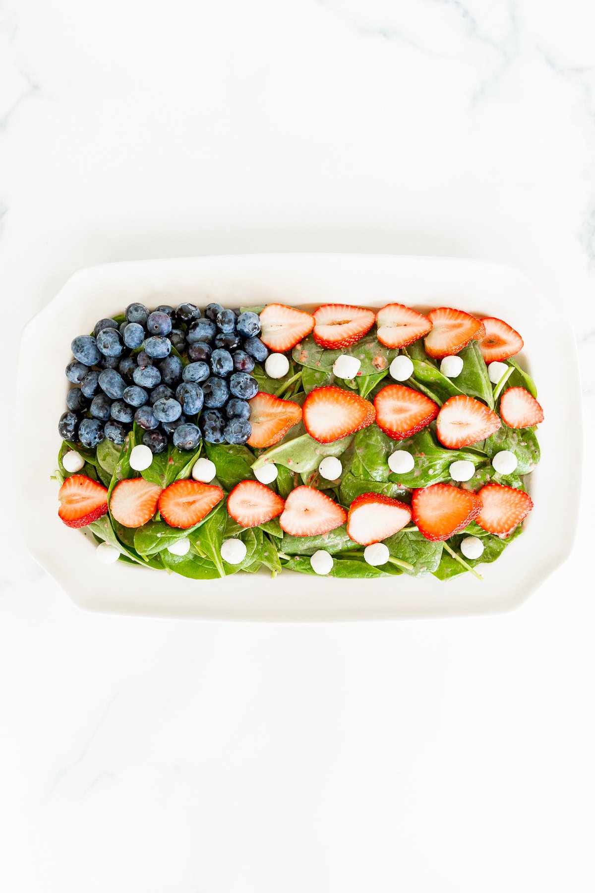 A rectangular platter with a red, white, and blue salad consisting of spinach topped with sliced strawberries, whole blueberries, and small white cheese pieces.