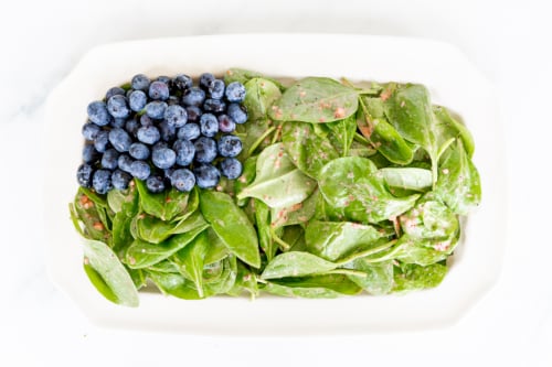 A white dish containing fresh spinach leaves topped with a dressing and blueberries