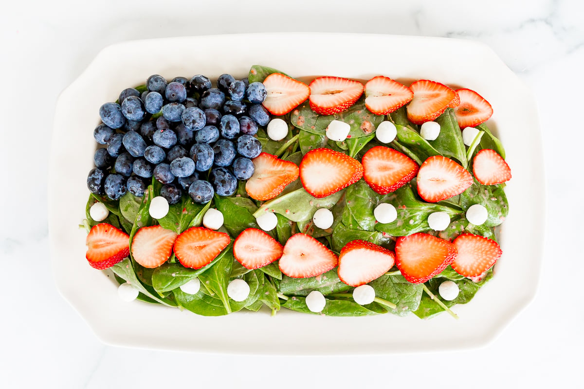 A red, white, and blue salad featuring spinach leaves, sliced strawberries, whole blueberries, and almond slices in a white dish on a marble surface.
