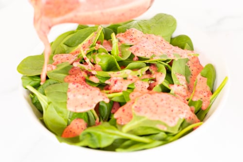 A jar of pink dressing pouring over a bowl of green spinach leaves.