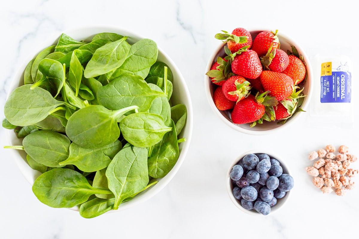 Overhead view of a bowl of fresh spinach, a bowl of strawberries, a smaller bowl of blueberries on a marble surface