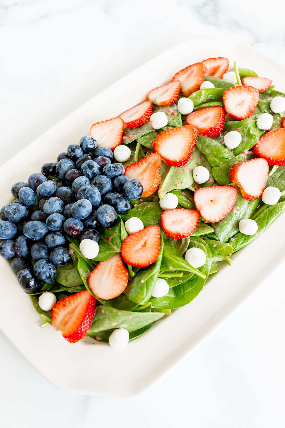 A vibrant red, white, and blue salad with fresh spinach, sliced strawberries, whole blueberries, and white round candies, all arranged in a white rectangular dish on a marble surface.