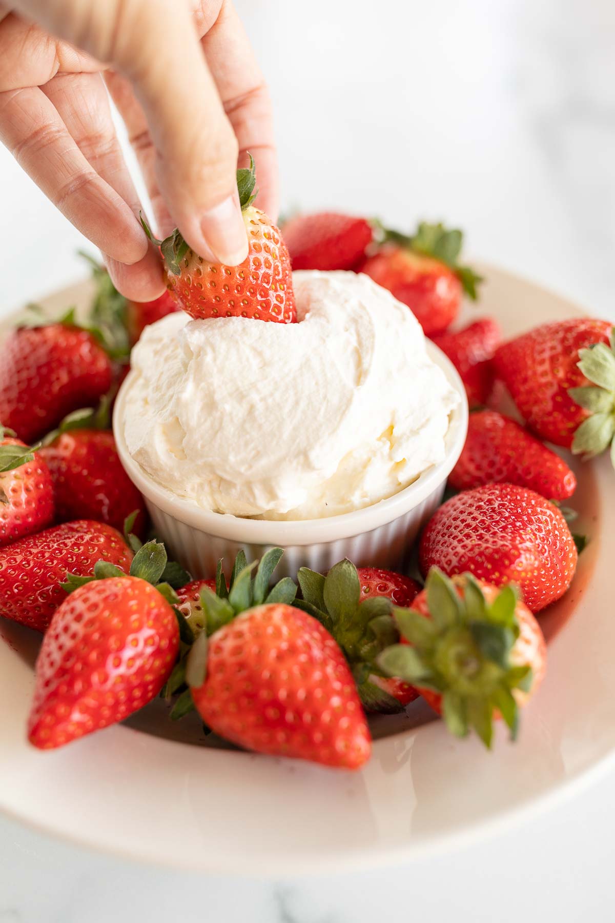 A plate of strawberries surrounding a bowl of fruit dip, a hand dips a strawberry into the dip.