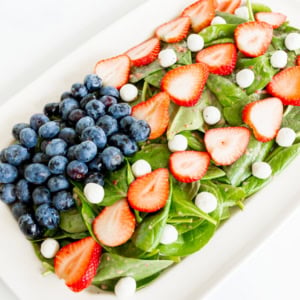 A fresh spinach salad topped with blueberries, sliced strawberries, and small white cheese balls, served as a patriotic red, white, and blue salad on a rectangular plate.