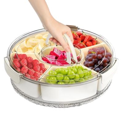 A hand lifts the clear lid of a circular serving tray divided into sections, much like snackle boxes, filled with assorted fresh fruits including grapes, strawberries, raspberries, bananas, and dragon fruit.