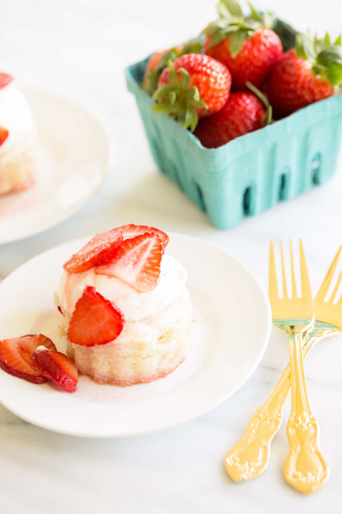 strawberry shortcake on a white plate, blue box of strawberries in background. 