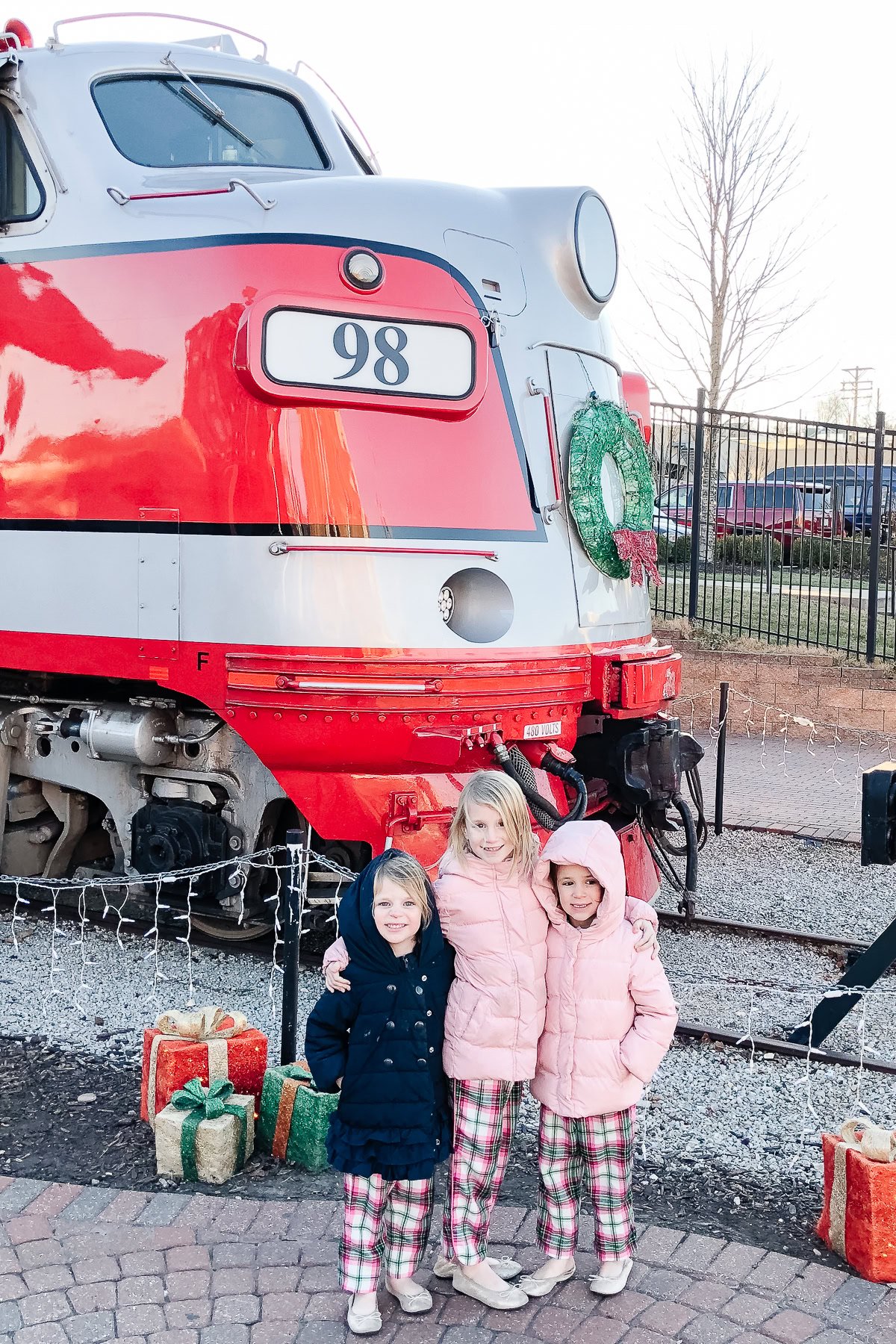 Three children in winter coats and matching plaid pants stand in front of a festive red and silver train engine decorated with a green wreath, one of the many charming things to do in Branson. Wrapped presents are placed on the ground nearby.