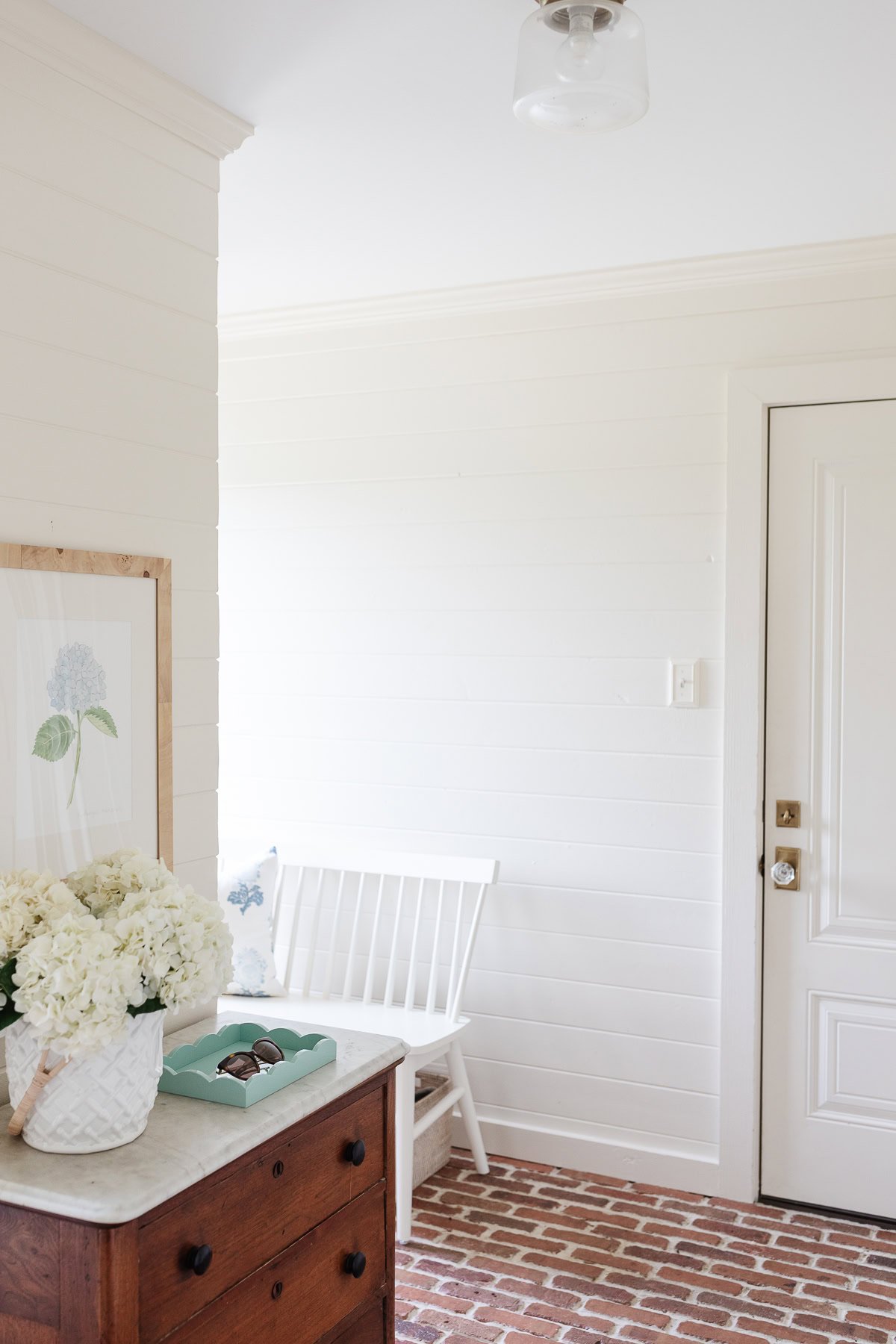 A bright entryway with thin brick pavers flooring, white walls, a white door, a wooden dresser with flowers, a framed botanical print, and a white bench.
