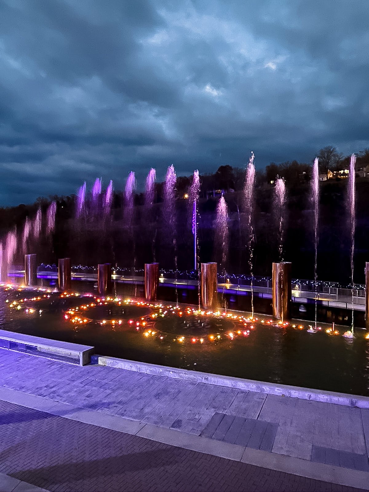 A nighttime scene of a water fountain with purple-lit jets and glowing lights in the pool, set against a cloudy sky—a must-see among things to do in Branson.