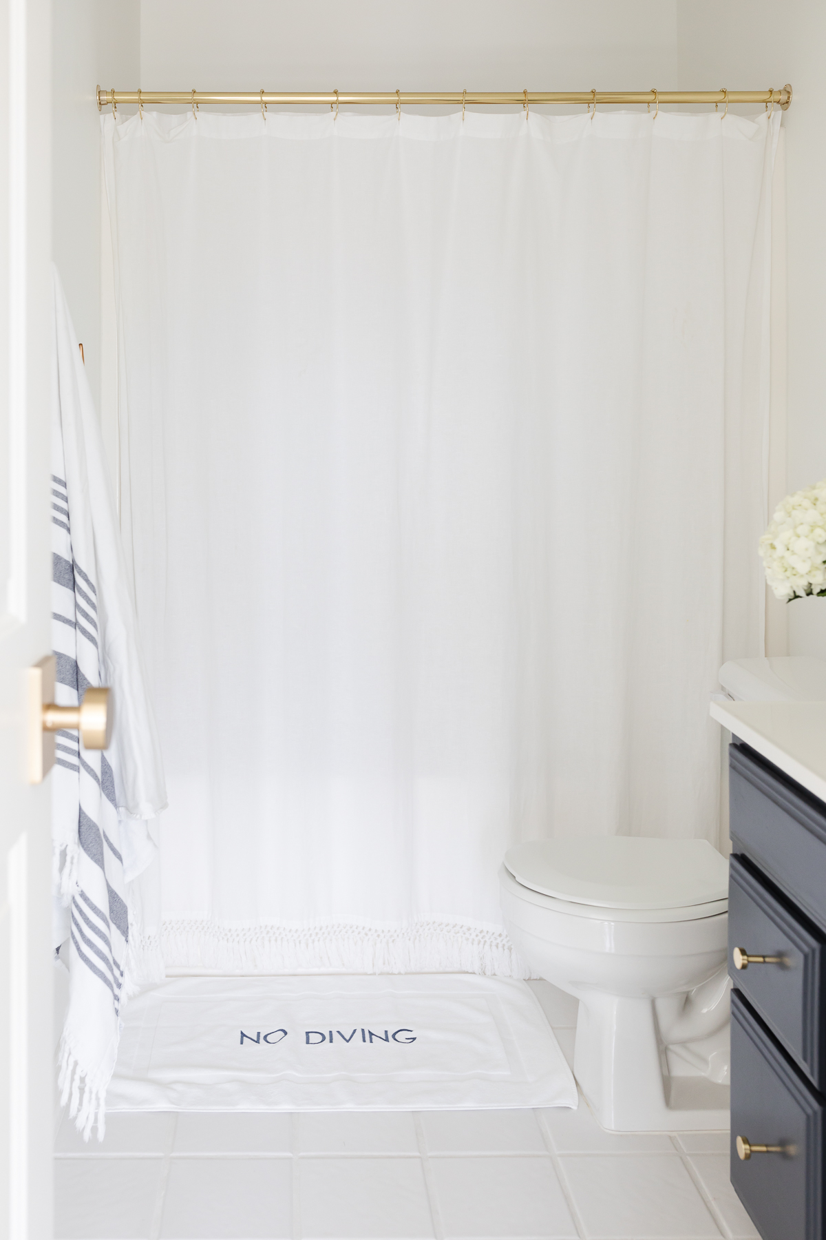 A white bathroom with a navy painted vanity and a gold shower curtain rod with a white tasseled curtain.