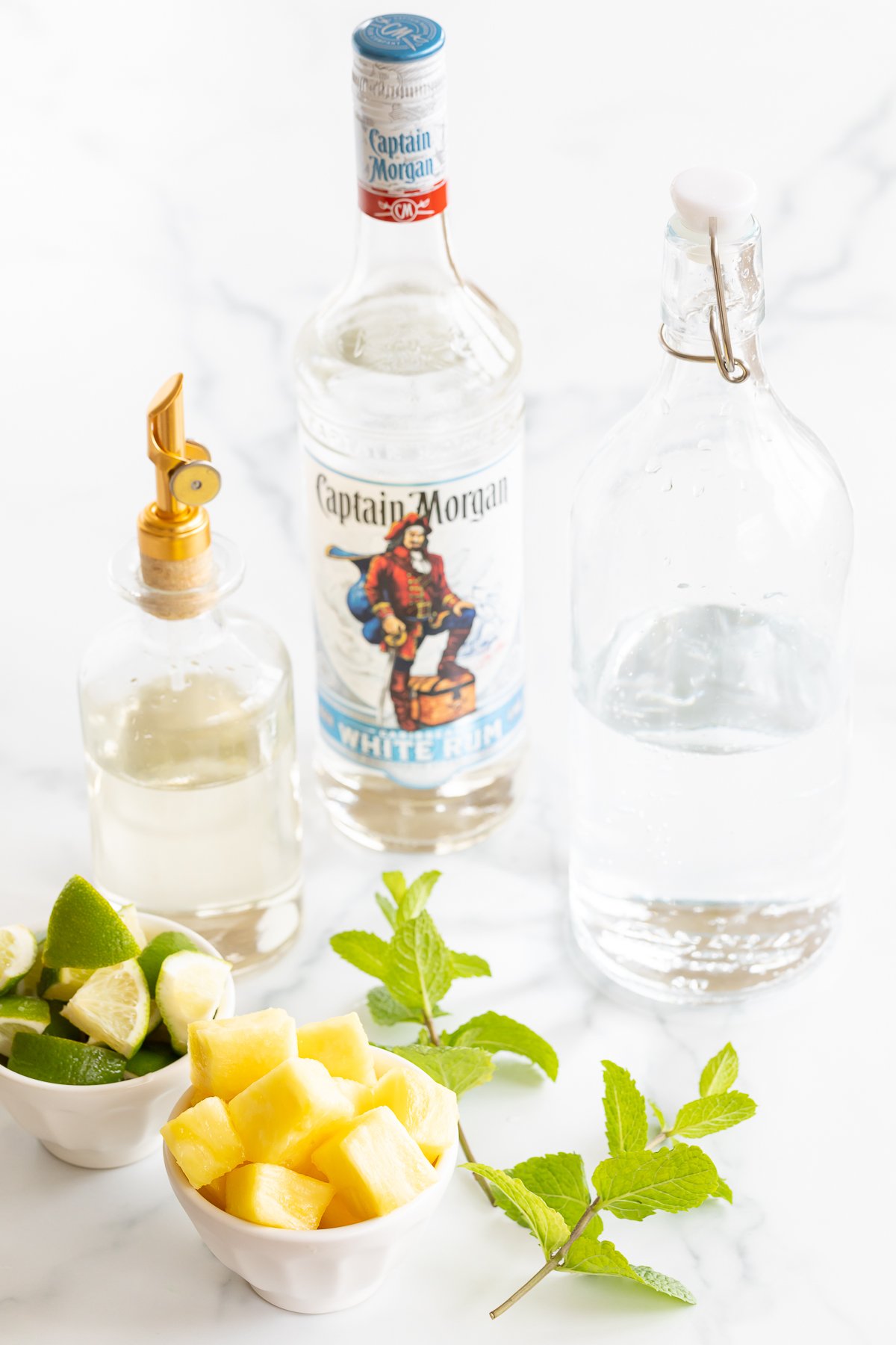 Bottle of pineapple simple syrup with bottles of white rum and club soda, bowls of lime wedges and pineapple, and sprigs of fresh mint