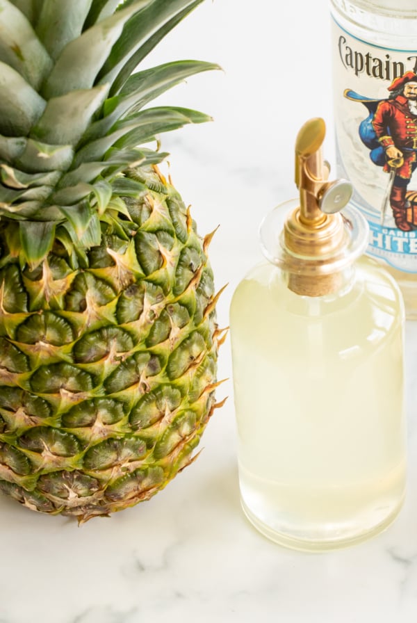 Bottle of pineapple simple syrup set next to fresh pineapple and bottle of white rum
