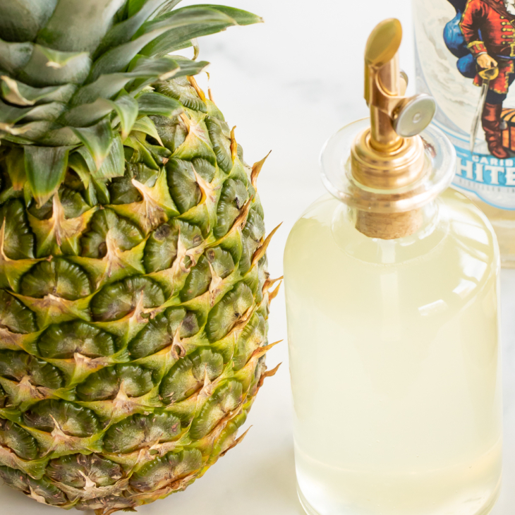 Bottle of pineapple simple syrup set next to fresh pineapple and bottle of white rum