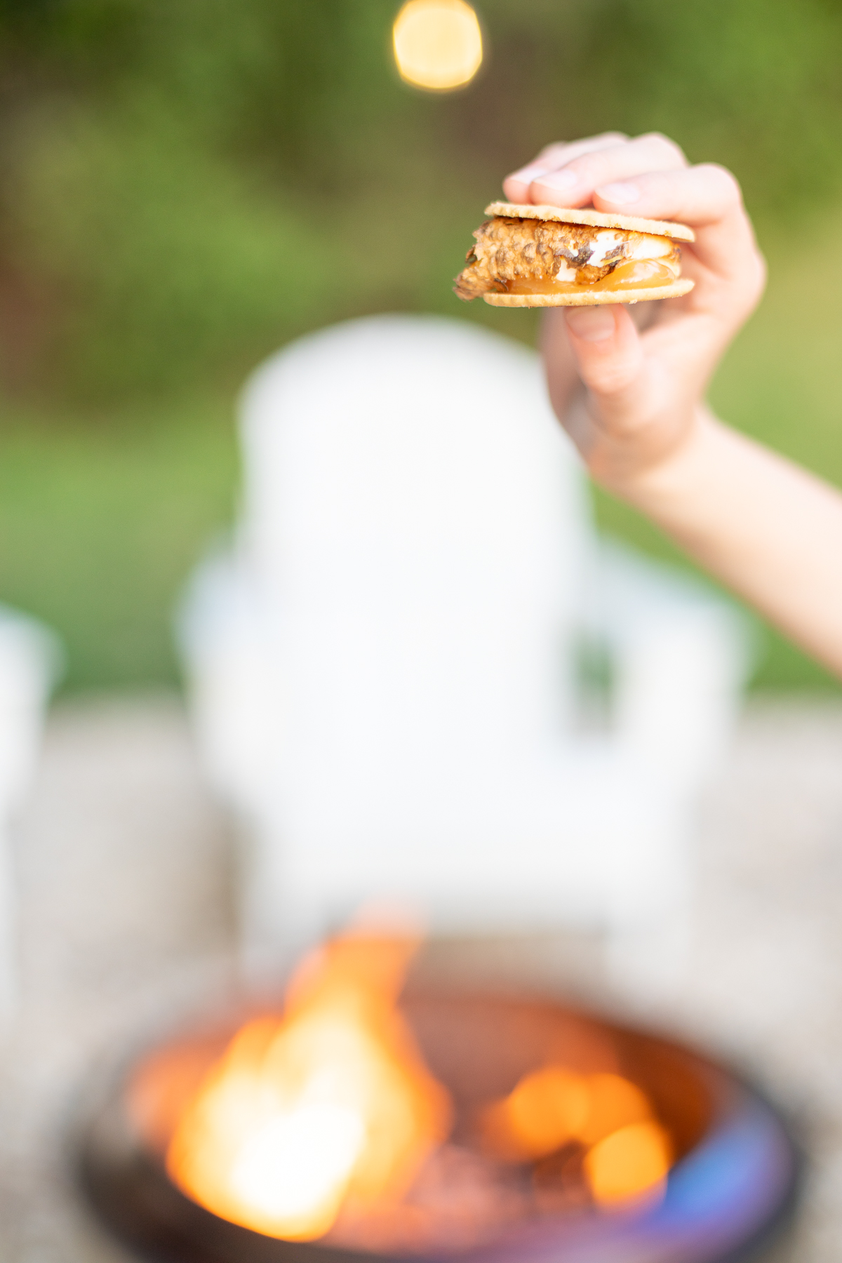 A hand holding a s'mores with a firepit in the background
