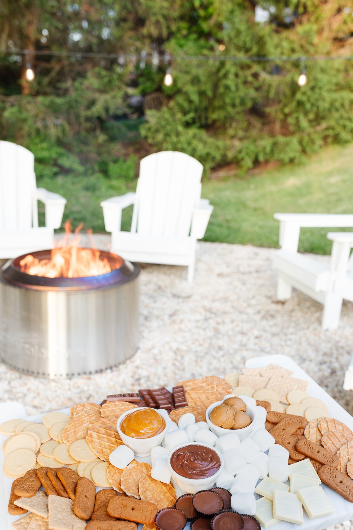 A Solo stove campfire on a gravel fire pit, surrounded by white chairs. A smores charcuterie board rests on one chair.