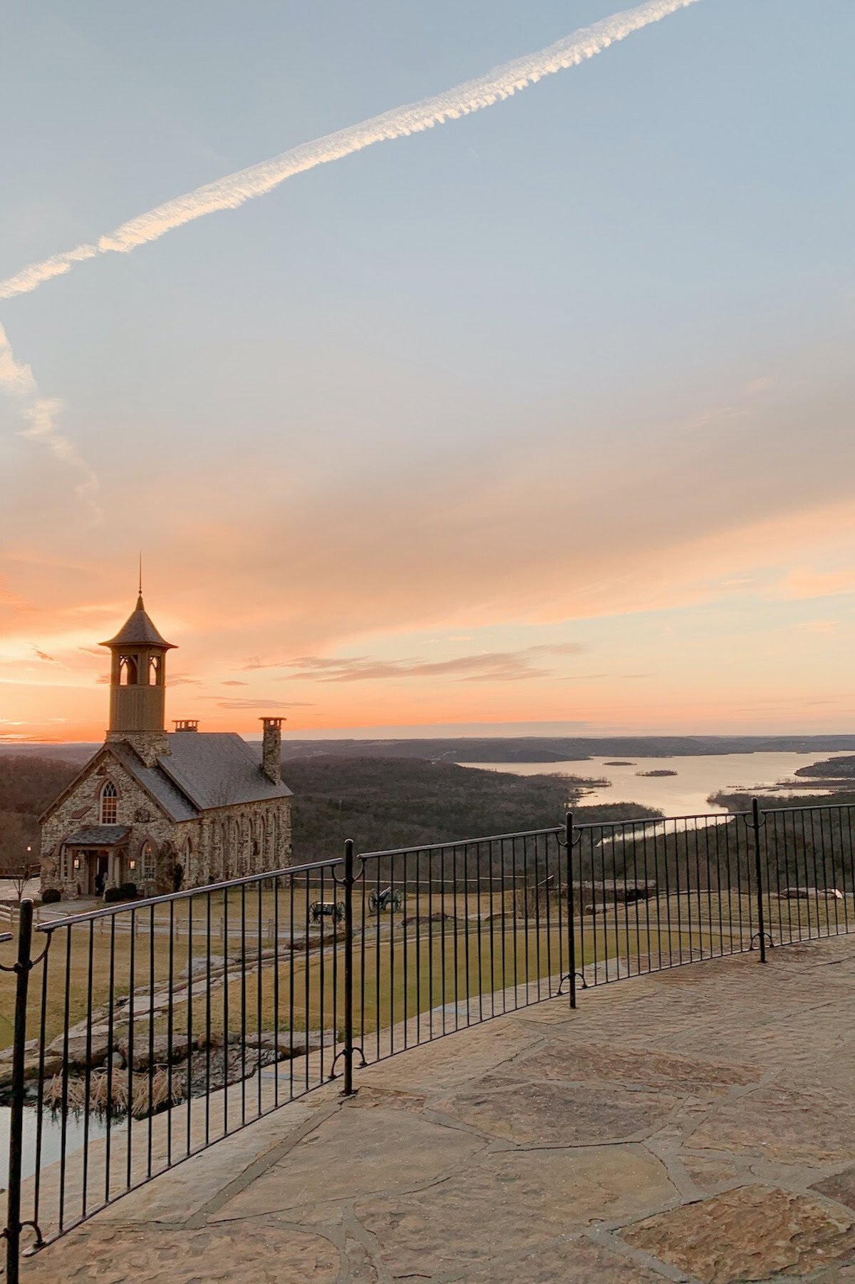 A stone church with a steeple at sunset overlooks a vast landscape of hills and a lake, bordered by a black iron fence in the foreground. It's one of the serene spots to visit among many things to do in Branson.