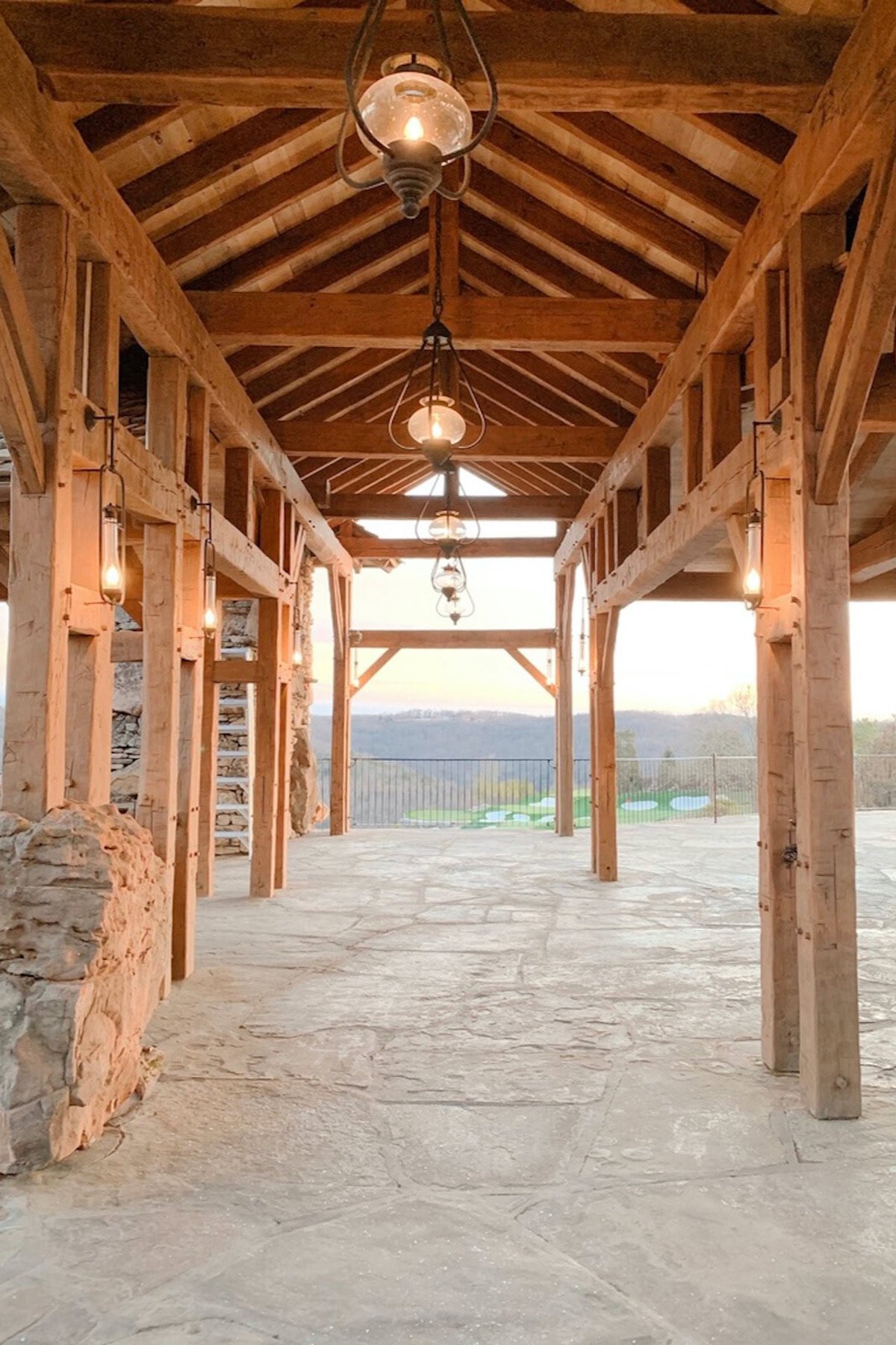 A covered walkway with wooden beams, hanging lanterns, and stone flooring overlooking a scenic landscape at sunset is one of the enchanting things to do in Branson.