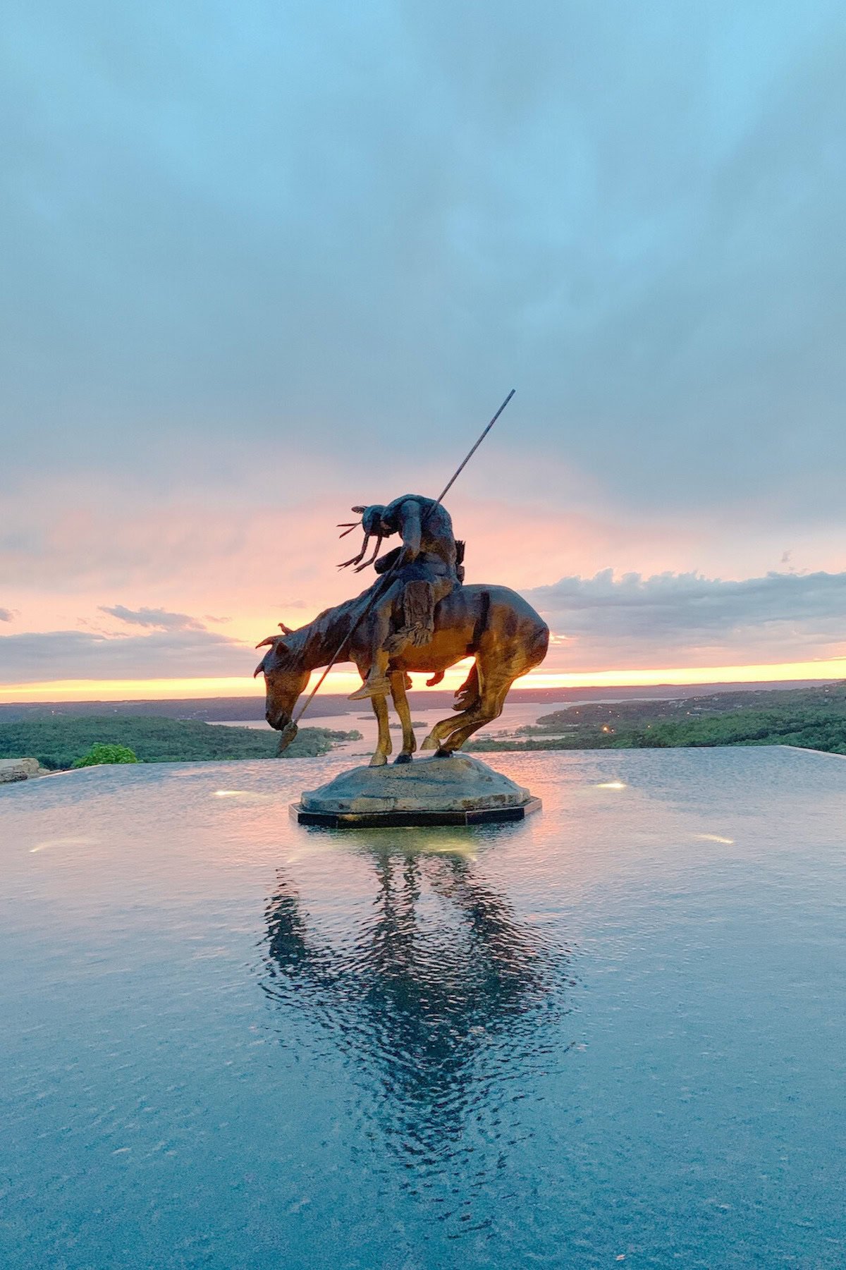 A bronze statue of a man on a horse, both appearing tired, stands on a small platform surrounded by water. This remarkable sight is one of the many things to do in Branson, where the background features a colorful sunset over a vast landscape.