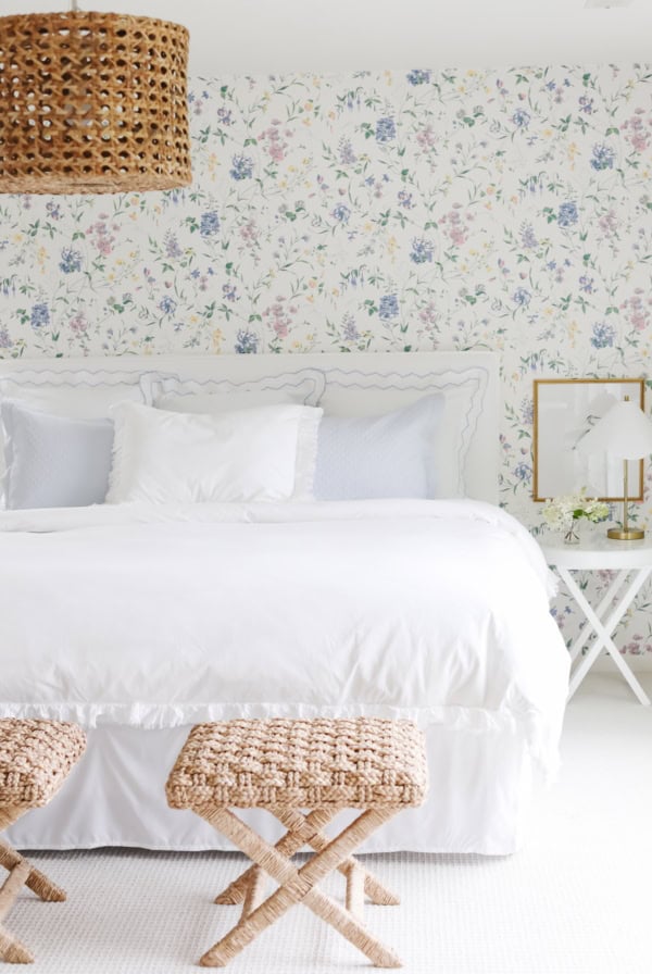 A bedroom with a white bed, Amazon Wallpaper featuring florals, two woven stools at the foot of the bed, a wicker pendant light, and a white side table with a lamp and flowers.