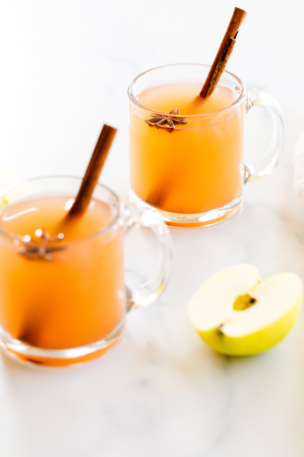 A marble surface with clear mugs full of homemade apple cider, whole apples to the side.