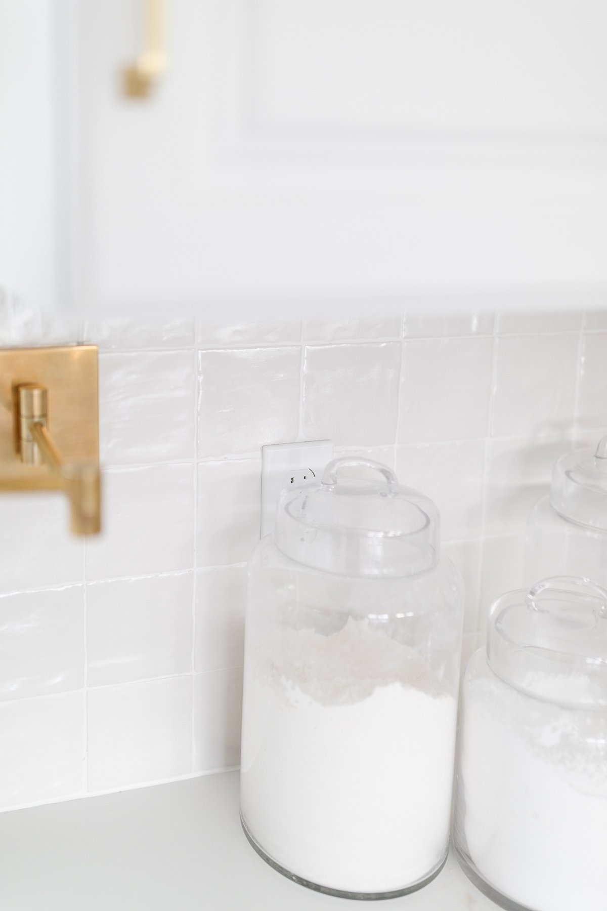 A glossy stacked tile backsplash in a white kitchen, glass canisters with baking supplies rest on the counter.