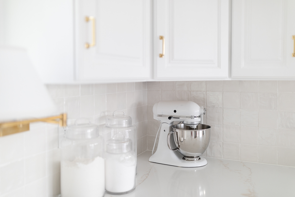 A glossy stacked tile backsplash in a white kitchen, glass canisters with baking supplies rest on the counter.