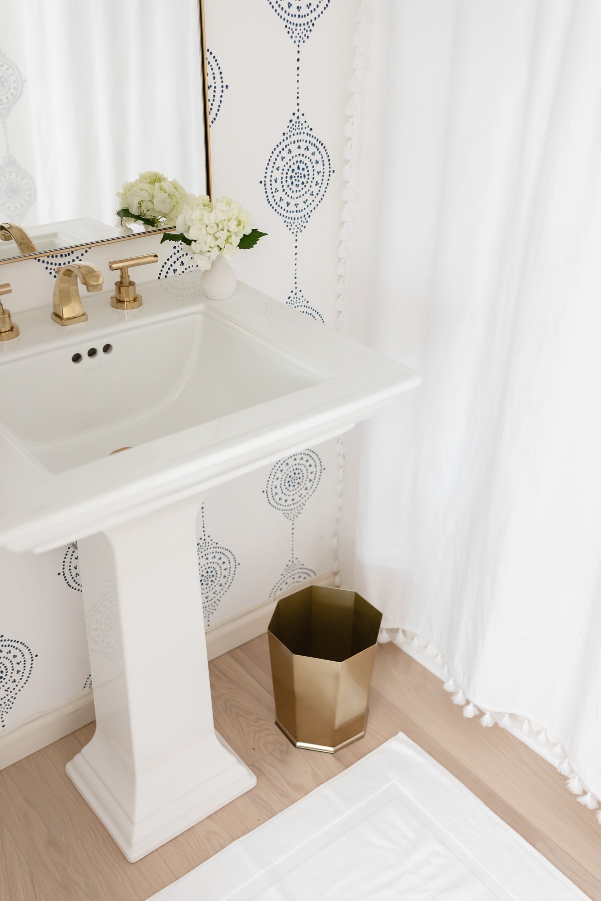 white bath with white and blue patterned wallpaper, pedestal sink and brass accents