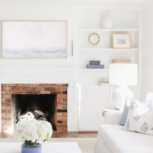 A living room with a white fireplace, a framed seascape painting above the mantel, built-in shelving, and a white sofa.