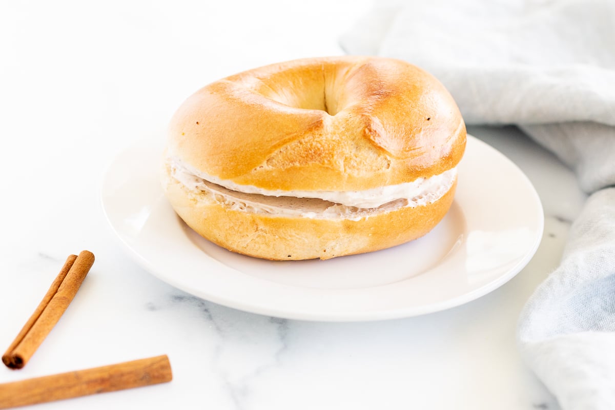 A bagel with cinnamon cream cheese, on a white plate with cinnamon sticks.