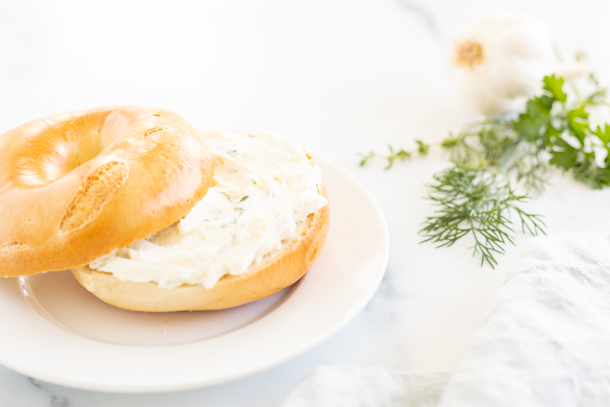 A bagel topped with garlic and herb cream cheese on a white plate.