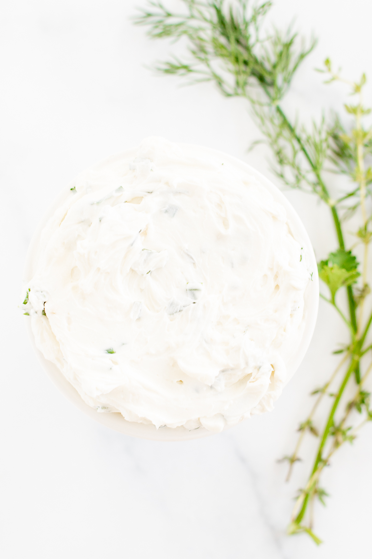A white bowl of garlic and herb cream cheese on a marble countertop. Fresh herbs nearby.