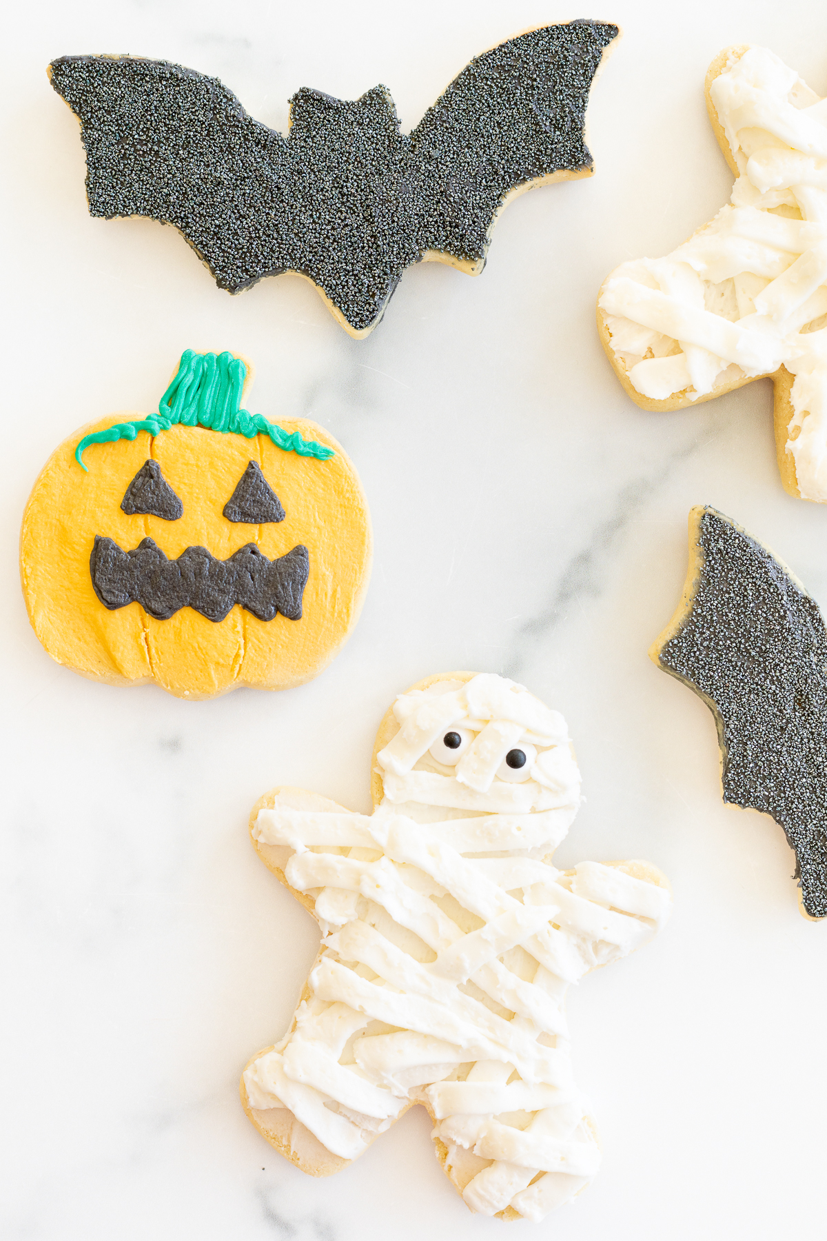 A variety of bat, jack o lantern and mummy Halloween sugar cookies laid out on a marble countertop.
