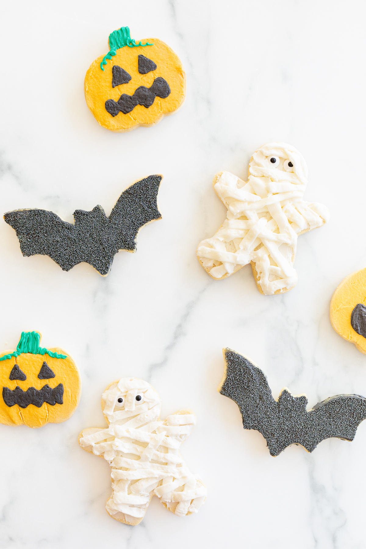 A variety of bat, jack o lantern and mummy Halloween sugar cookies laid out on a marble countertop.