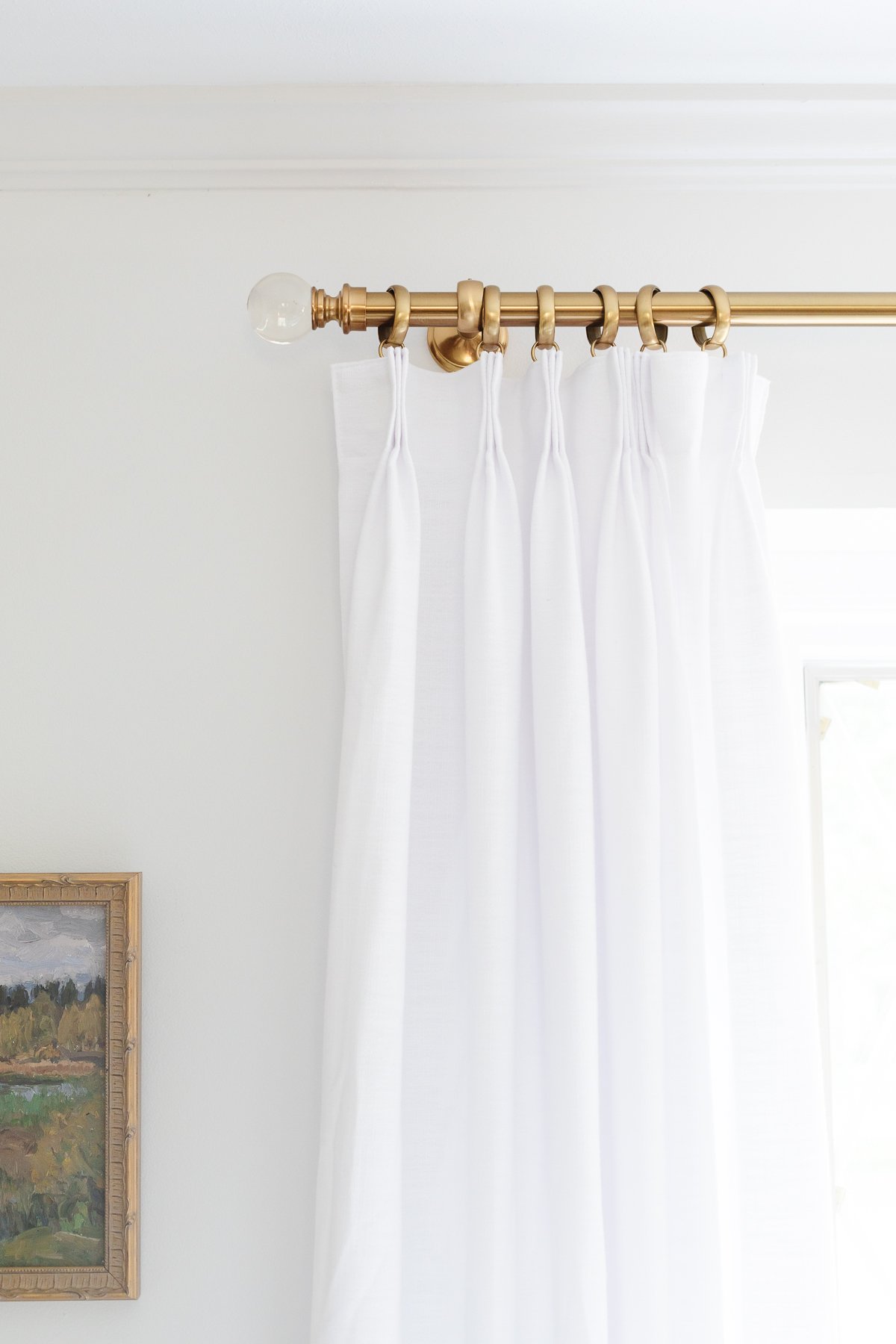 A brass curtain rod with white triple pinch pleat drapes in a living room.