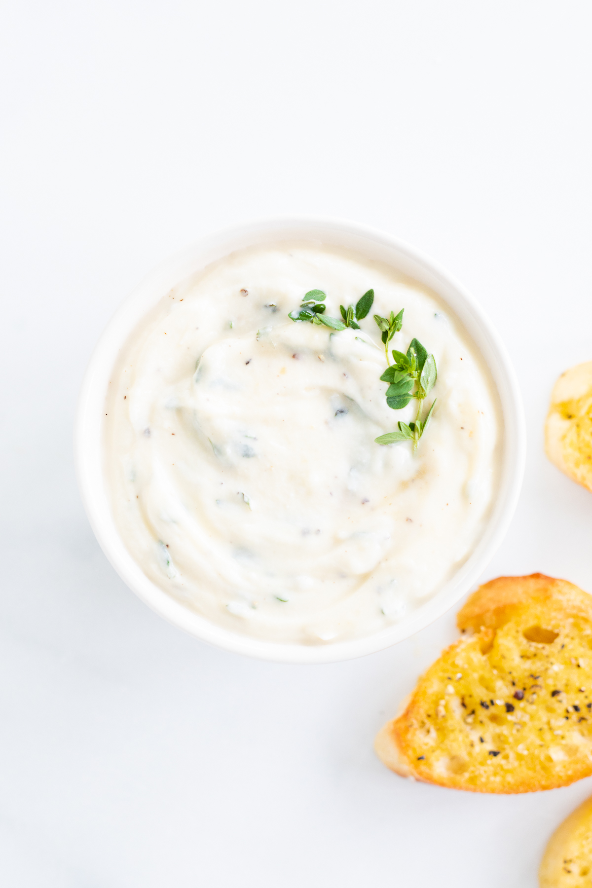 A bowl of whipped ricotta dip with bread and herbs on a white background.