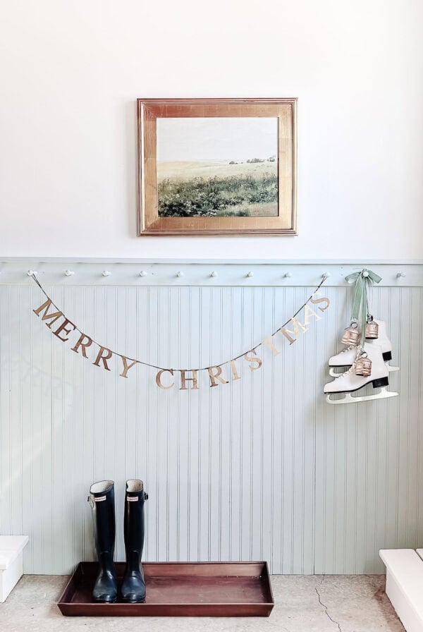 A gold "Merry Christmas" garland and shiny brass bells hang on a pale blue wall; black boots and a tray rest below, with white ice skates nearby and a framed landscape painting above.