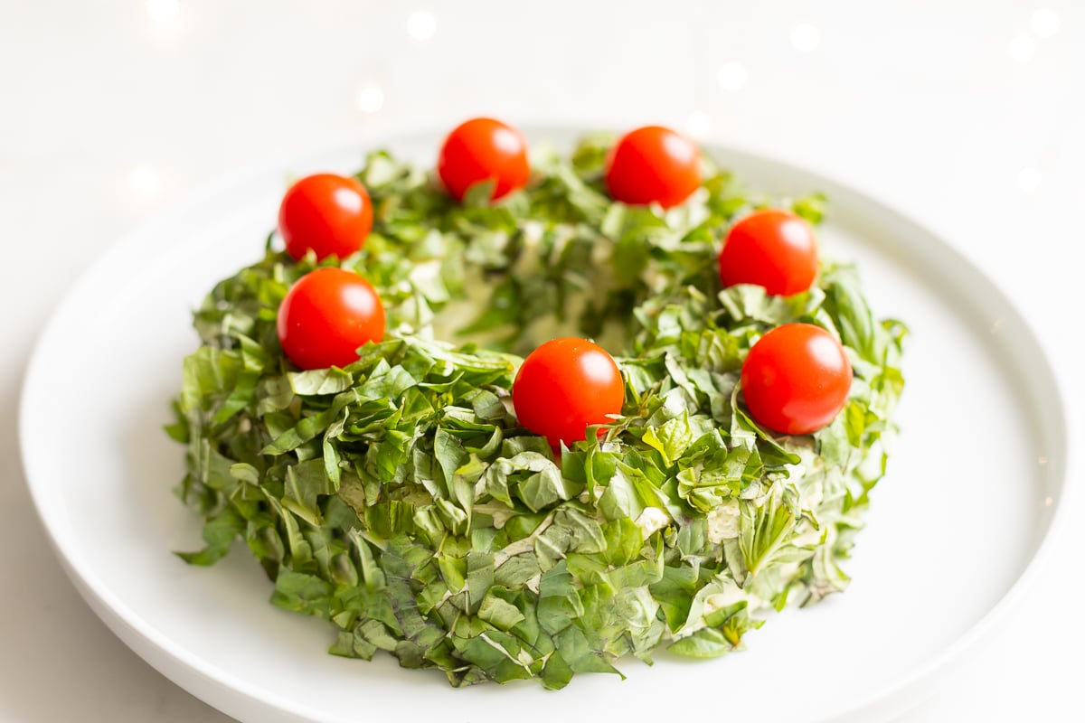 A plate with a Christmas cheese ball wreath decorated with basil and tomatoes.
