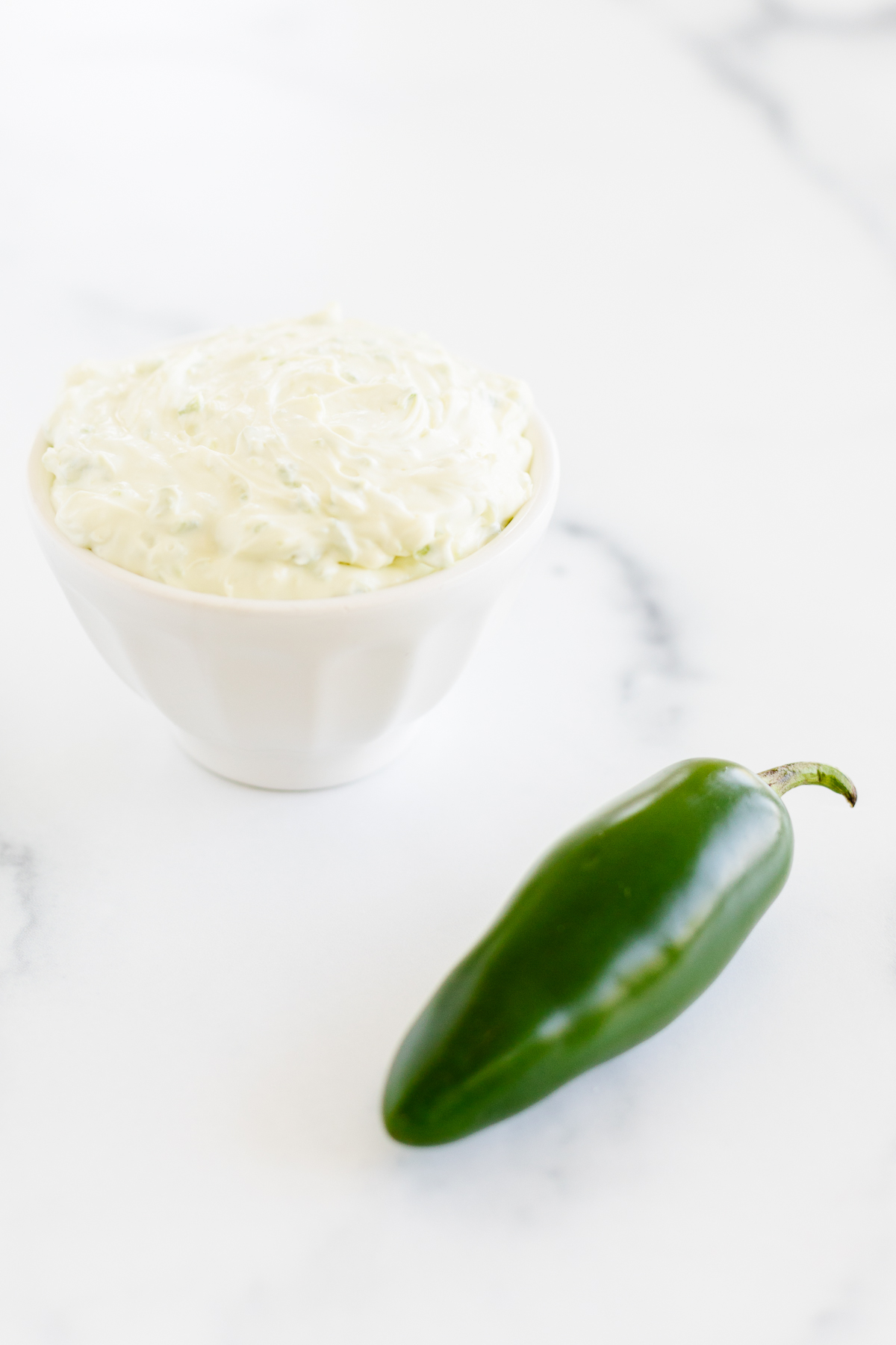 A white bowl with a jalapeno and a green pepper, alongside a dollop of cream cheese.