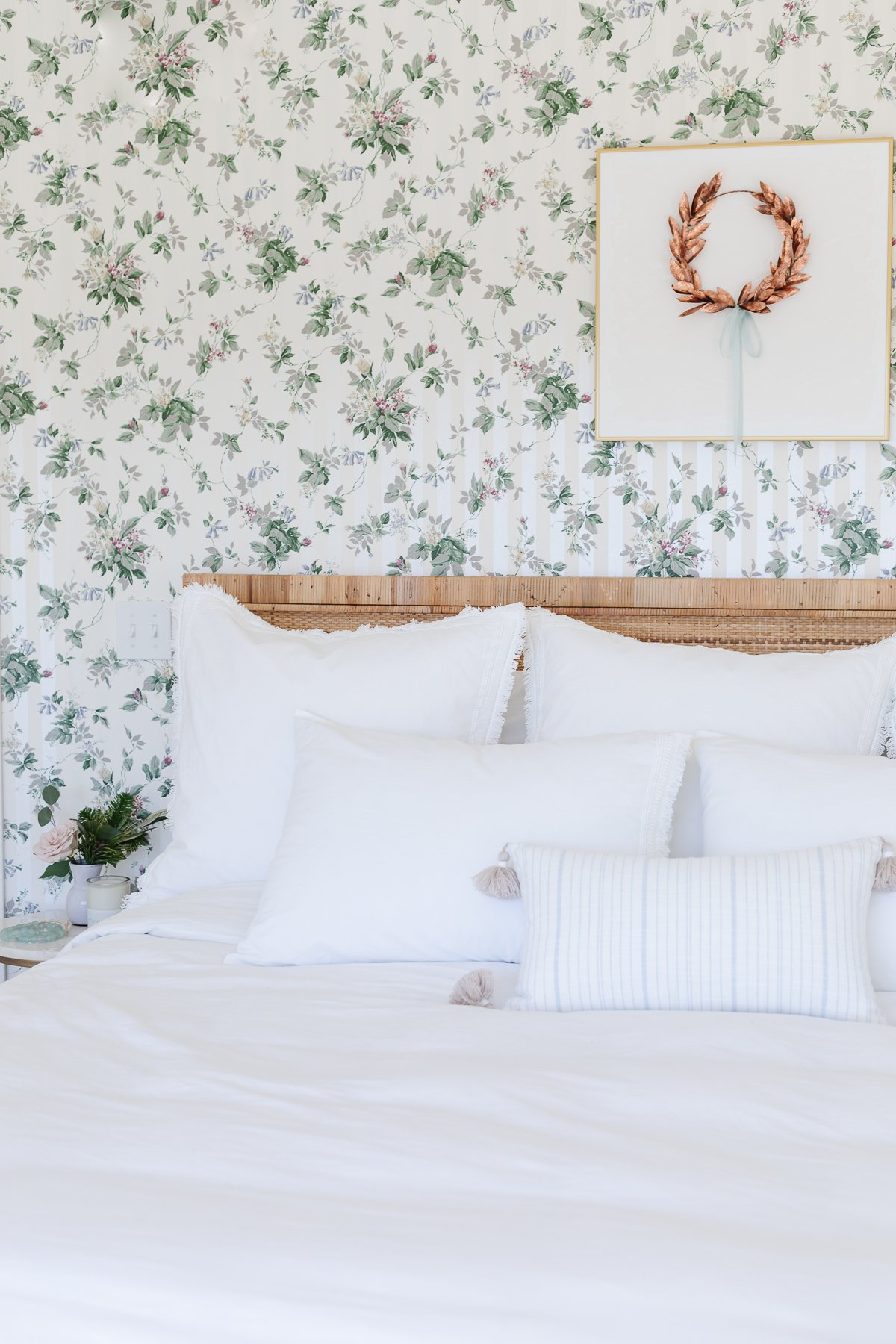 A white bed in a bedroom that has floral wallpaper, with a gold laurel wreath hanging over the white art above.