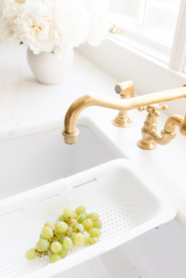 A kitchen sink adorned with unlacquered brass faucets, complemented by clusters of grapes immersed in water.