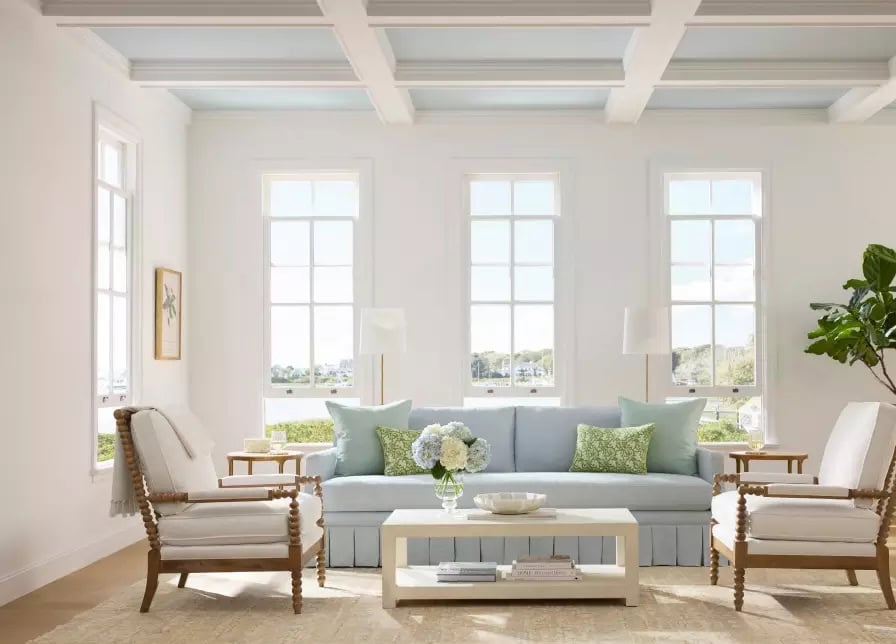 A bright living room with a Serena and Lily Sofa in light blue, two wooden armchairs, a coffee table with flowers, and large windows letting in natural light.