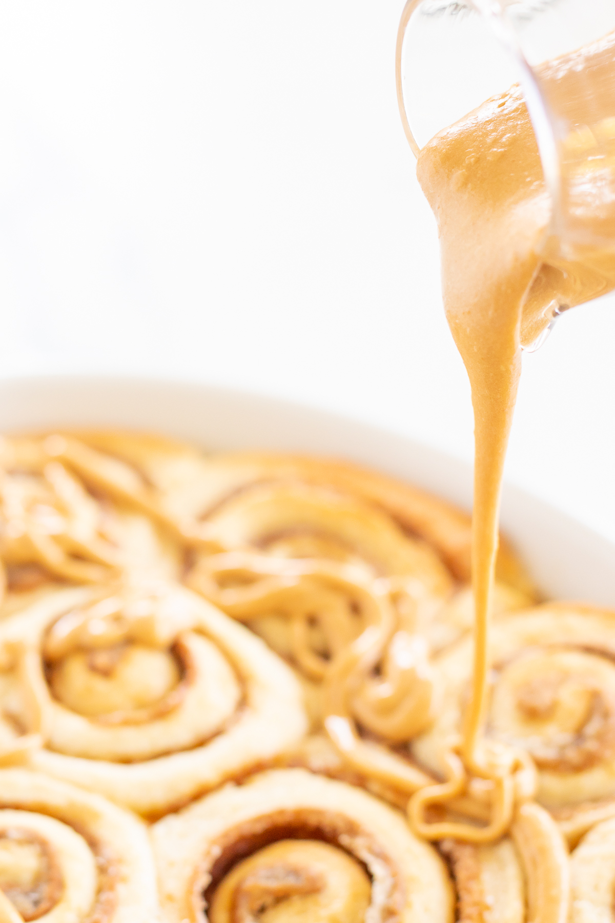 A clear glass pitcher pouring cookie butter frosting over a pan of cinnamon rolls.