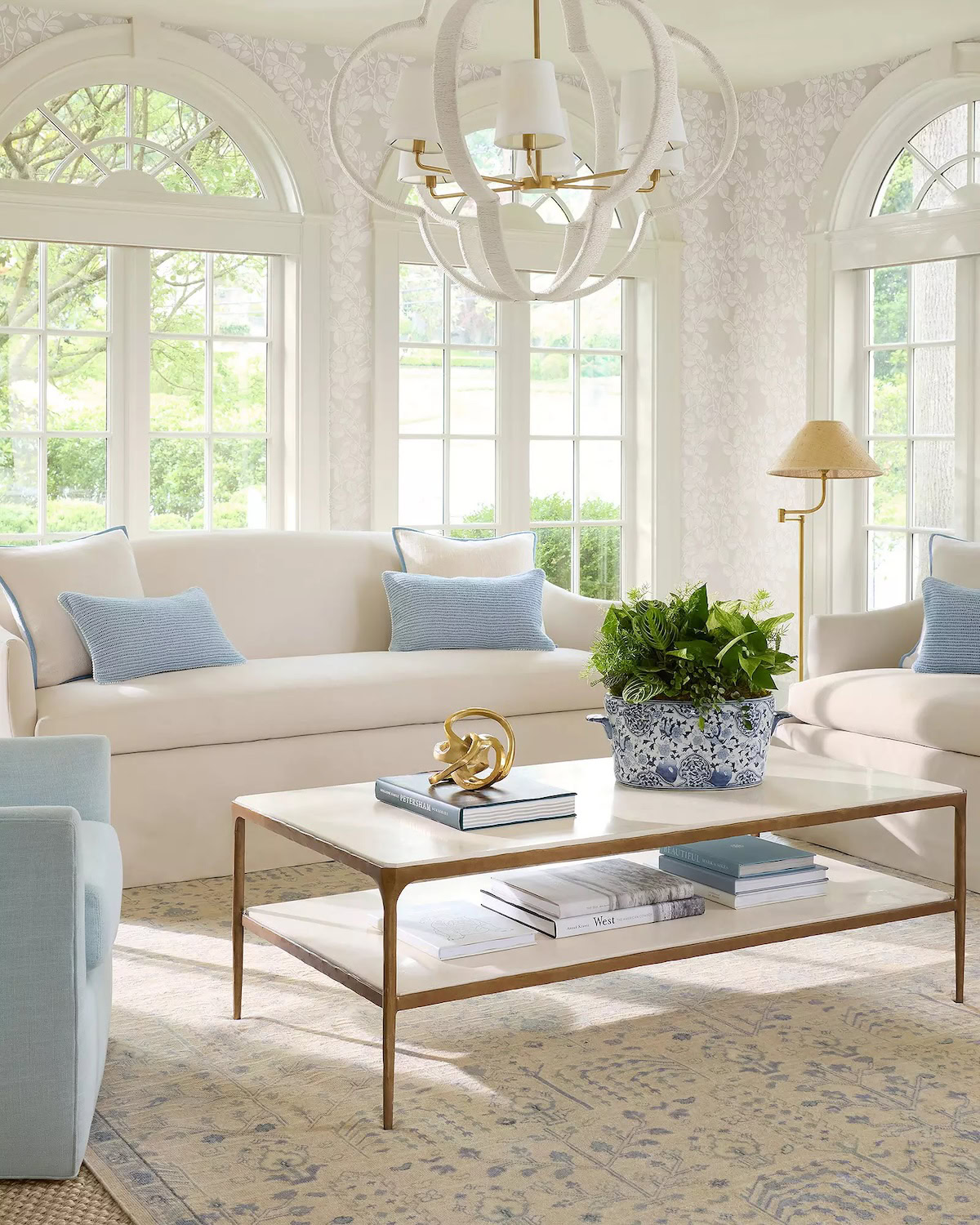 A bright living room with two Serena and Lily sofas, light blue pillows, a modern chandelier, large windows, and a coffee table adorned with books, a plant, and a decorative object.