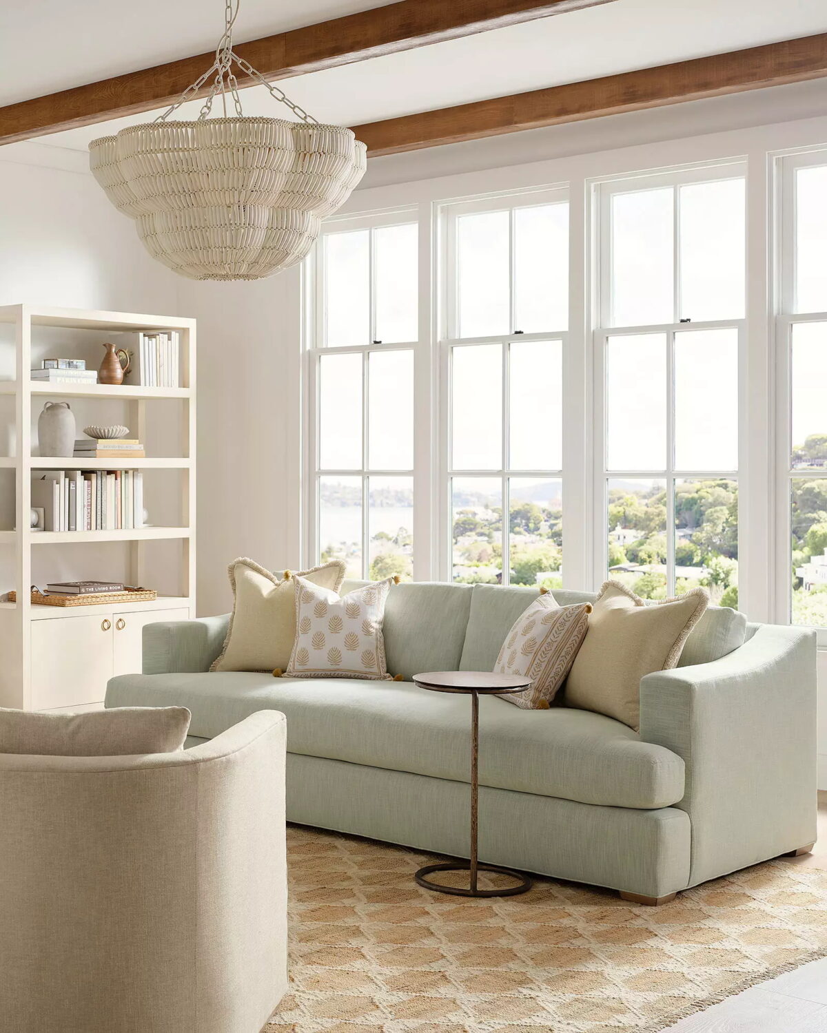 A light-filled living room with a Serena and Lily Sofa in pale green, neutral pillows, a round side table, a bookcase, and large windows overlooking a scenic view.