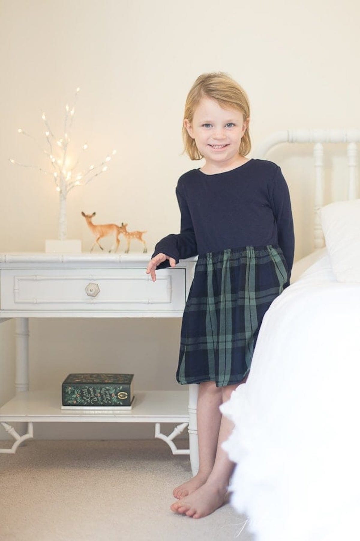 A little girl in a plaid dress standing in front of a white dresser, filled with Christmas magic.