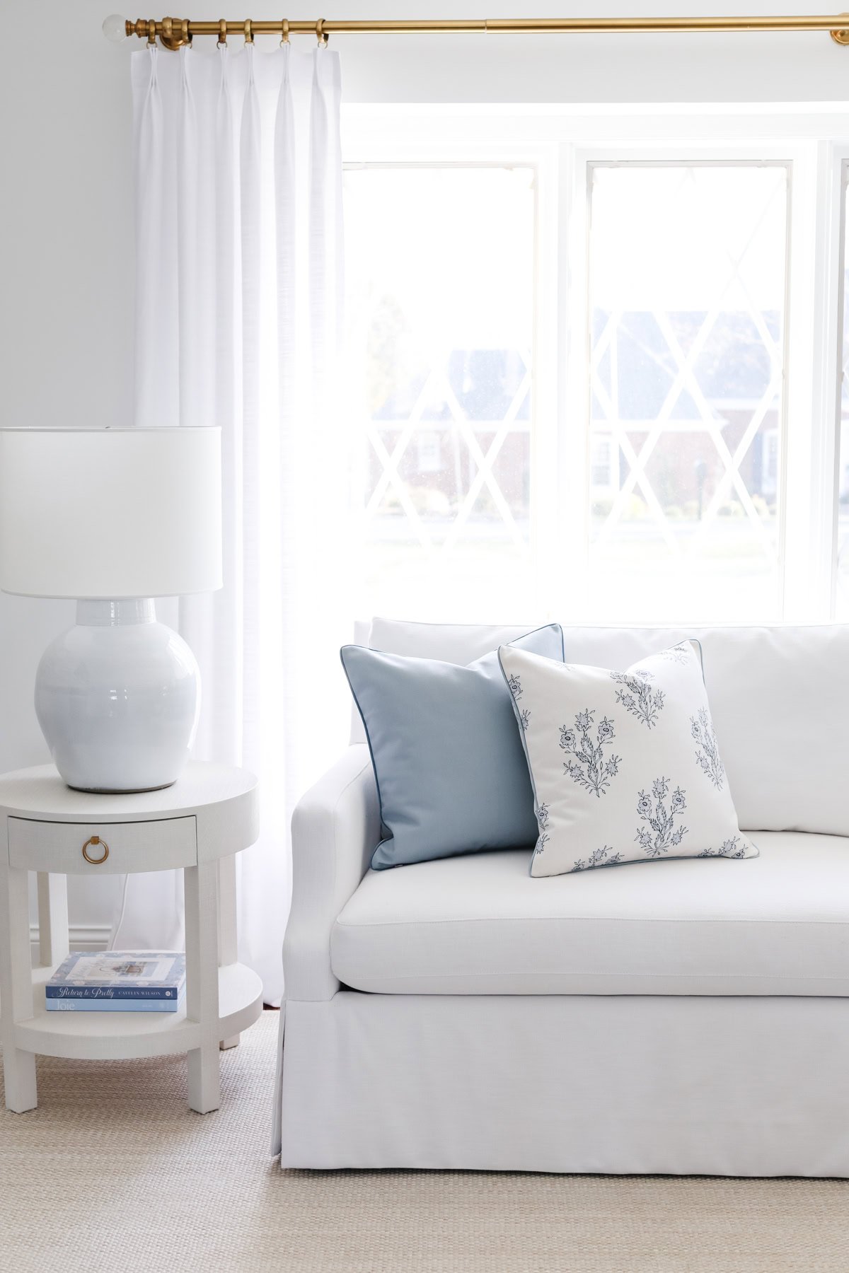 A white sofa with two decorative pillows, one light blue and one white with blue patterns, is placed against a window. Achieve the RH Look for Less with a matching white side table and lamp sitting next to it.