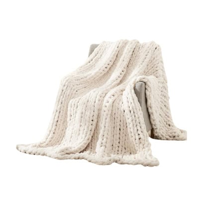 A cream-colored chunky knit blanket, giving off a Pottery Barn look for less, is draped over the arm of a grey chair.