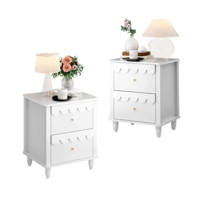 Two white bedside tables with two drawers each exude a Pottery Barn Kids look for less. One boasts a lamp, flowers, and small decor items; the other features a lamp, books, and a candle. Both have round knobs and scalloped edges on the drawers.