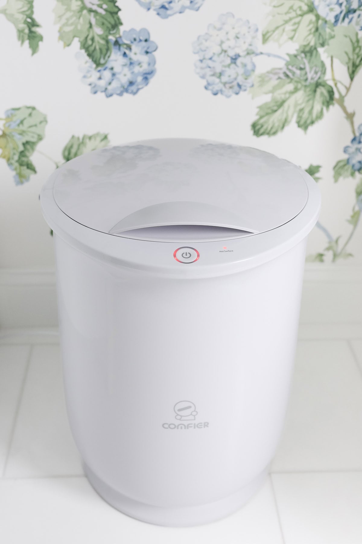 A white countertop compost bin with a lid and power button—one of the latest must-have Amazon gadgets—sits on a tiled floor in front of floral wallpaper.
