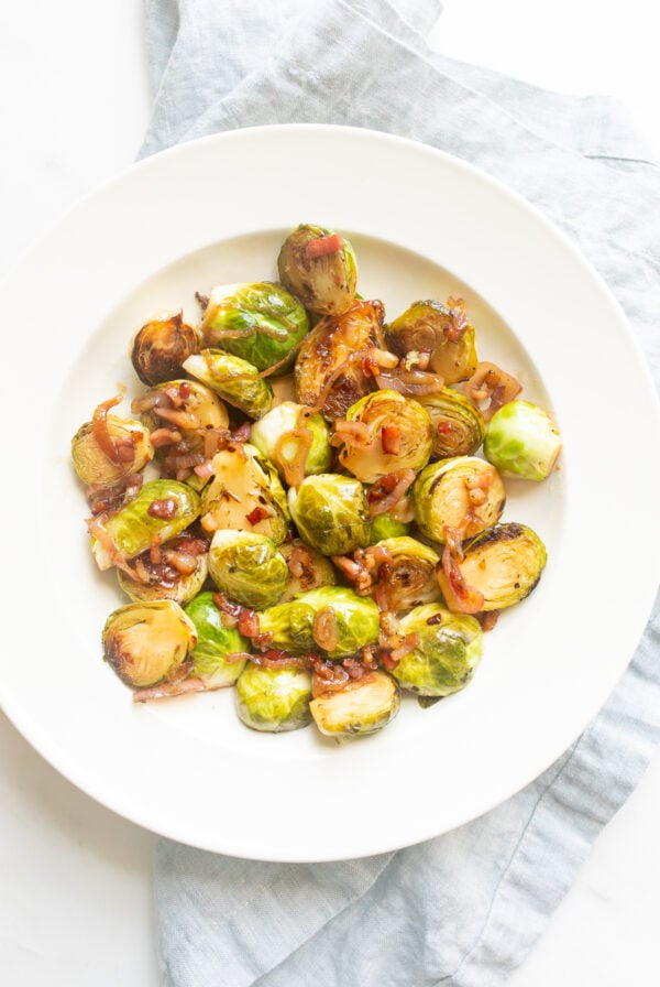 Crispy brussel sprouts on a white plate with a blue linen napkin and serving spoon to the side.